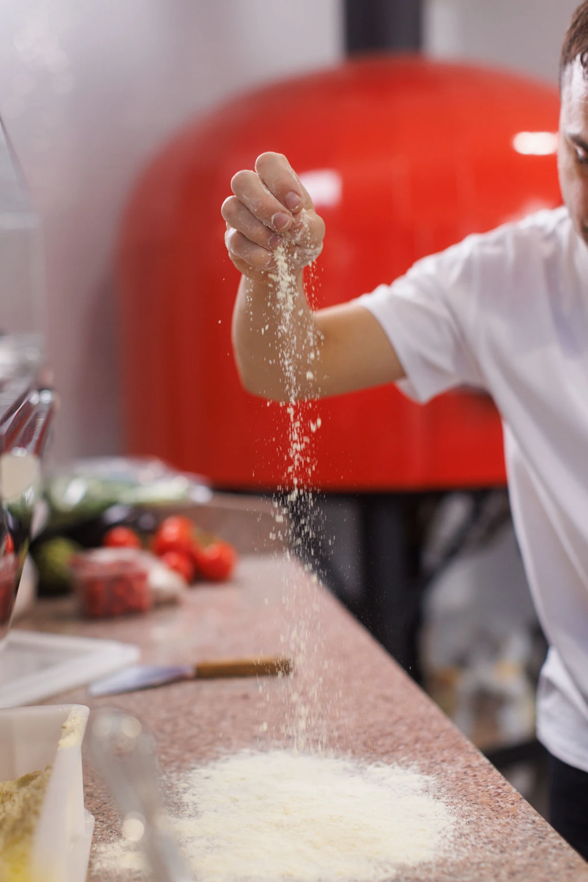 A pizza chef sprinkling flour onto a stone counter, with a red wood-fired oven and fresh tomatoes in the background.