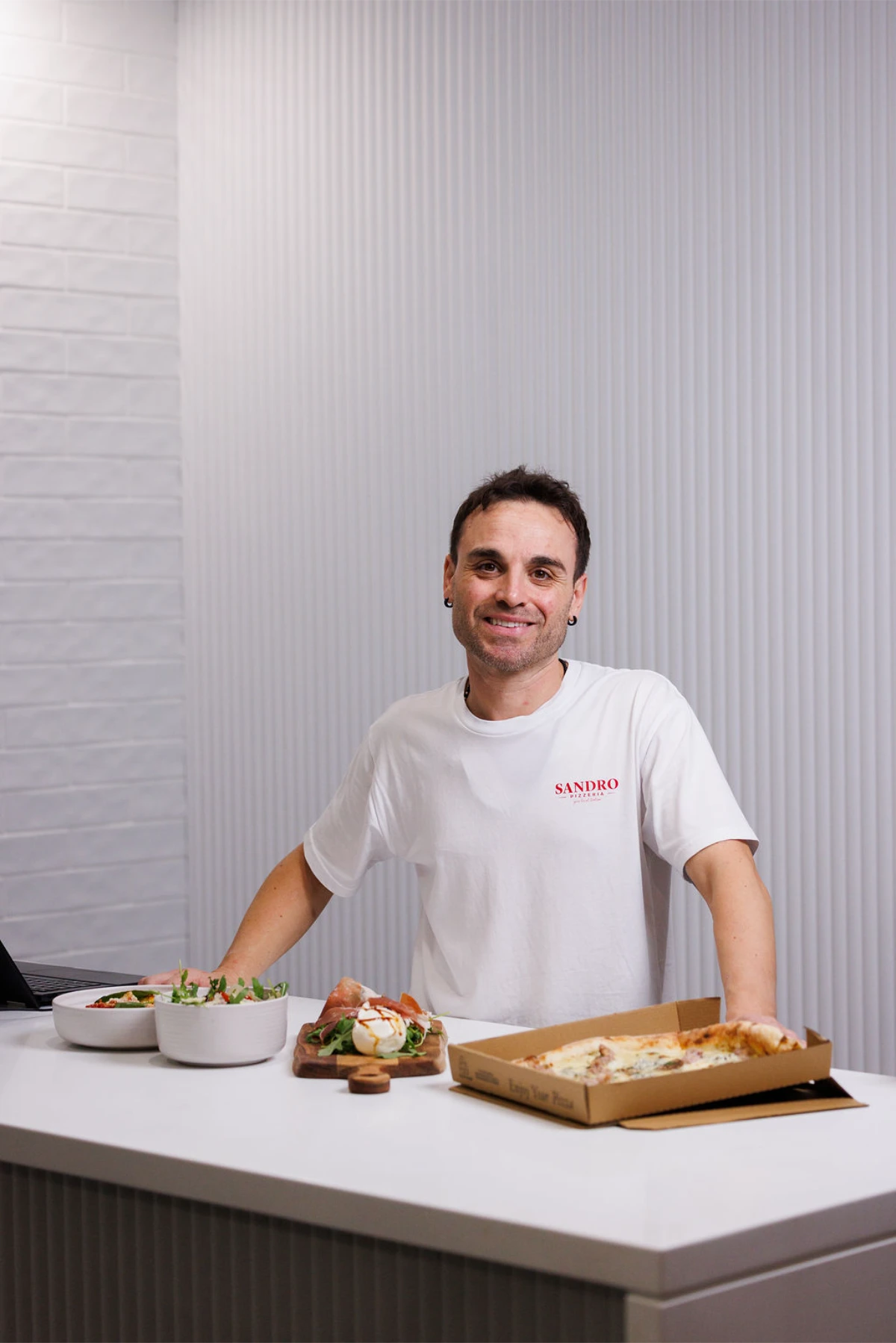 A smiling staff member in a Sandro Pizzeria t-shirt standing behind a counter with a boxed pizza, salads, and a burrata board.